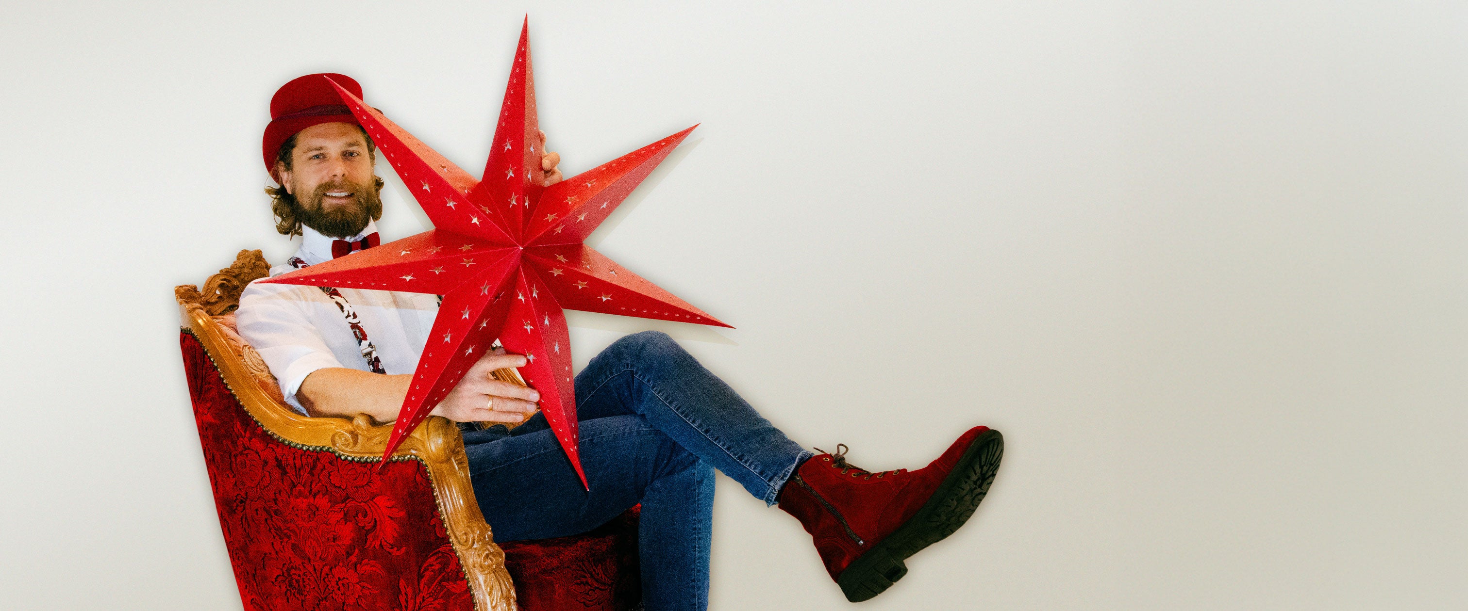 Man in white shirt, red hat, and boots, seated in a red armchair, holding a big red Christmas star